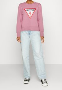 Sweat-shirt rose avec un grand logo triangulaire rouge portant le texte "GUESS U.S.A." sur la poitrine, associé à un jean bleu clair et des baskets noires.