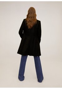 Abrigo negro de corte entallado, con cintura ceñida y mangas largas, combinado con jeans de mezclilla azul de pierna ancha. Textura suave.