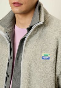 Fleecejacke in Hellgrau mit weicher Textur, ausgestattet mit einem grauen Kragen, durchgehendem Reißverschluss und einem bunten Logo-Patch auf der Brust.