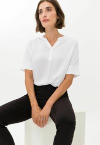 Weiße Bluse mit kurzen Ärmeln, V-Ausschnitt und Knopfdetail, kombiniert mit schwarzen, taillierten Hosen. Glattes Material, lässige Passform, minimalistisches Design.