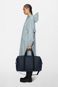 Torbica za prtljagu tamnoplave boje, izrađena od izdržljivog materijala, s crnim ručkama i podesivim remenom. Model nosi svijetloplati kišni ogrtač i čizme.