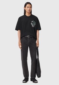Μαύρο βαμβακερό t-shirt με κοντά μανίκια και γραφικό τύπωμα στο αριστερό στήθος, συνδυασμένο με μαύρες τζιν παντελόνες και μαύρη τσάντα ώμου.
