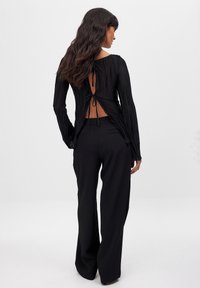 Black pleated long-sleeve top with open back design, tied at the waist, paired with loose-fitting black trousers. Smooth texture.