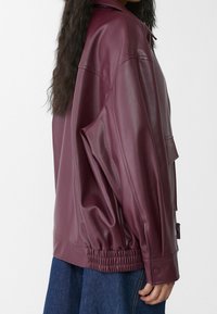 Burgundy konstläderjacka med en avslappnad silhuett, sidofickor och elastiska ärmslut. Slät textur och detalj med dragkedja.