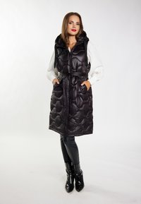 Gilet imbottito nero con motivo trapuntato, cappuccio e cintura, dotato di chiusura con zip e tasche laterali. Indossato sopra una maglietta bianca e pantaloni neri.