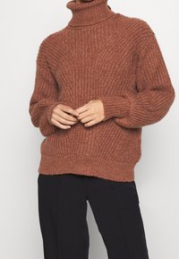 Raskumppuja neulepusero lämpimässä ruskossa, jossa on ribbikuvio, pudotetut olkapäät ja rento malli. Yhdistetty mustiin housuihin.