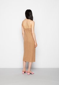 Nainen, jolla on pitkä musta hius, pukeutuneena hihattomaan beigeen neule-midi-mekkoon ja vaaleanpunaiseen matalan korokkeen liukuvaan tyyliin, seisoo valkoista seinää vasten.