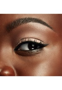 Närbild av ett öga med mörkbrun iris, definierad eyeliner, långa svarta ögonfransar och slät, bronsfärgad ögonlockshud. Tydlig detalj på ögonbrynets form.