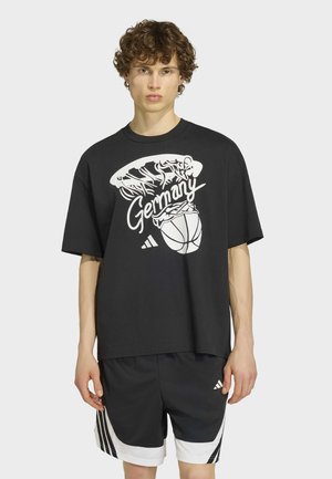 Junger Mann mit lockigem Haar trägt ein schwarzes übergroßes T-Shirt mit Basketball- und "Germany"-Aufdruck, kombiniert mit schwarzen und weißen Adidas-Shorts.