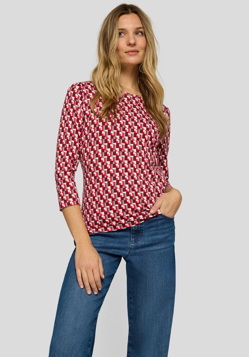 Rotes Oberteil mit geometrischem Muster in Weiß und Blau, aus einem weichen Stoff, kombiniert mit blauen Jeans.