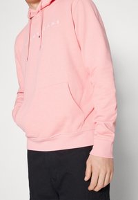 Osoba oblečená ve světle růžové mikině Tommy Jeans s přední kapsou a černými kalhotami, stojící proti jednoduchému světlem pozadí.