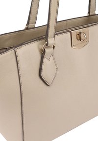 Sac fourre-tout en cuir beige avec un design structuré, deux poignées, quincaillerie dorée et un accent de tag décoratif. Texture lisse avec des détails de coutures.