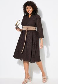 Braunes, knielanges Kleid mit Lochstickerei, Dreiviertelärmeln und einem Stoffgürtel. Getragen mit beigen Sandalen mit dicken Absätzen.