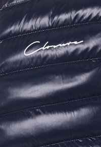 Glansigt marinblått kviltat tyg med vit snirklig "Clonwave"-logotyp sydd över materialet i ett horisontellt mönster.