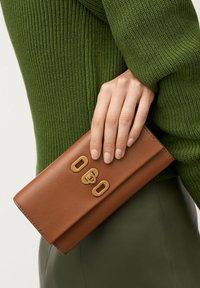 Portefeuille en cuir marron avec un design plié, mettant en avant des détails en métal doré et des gravures. Fini texturé et lisse. Tenue dans une main sur fond vert.