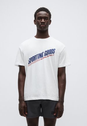 Maglietta bianca a maniche corte con stampa grafica blu e rossa con la scritta "SPORTING GOODS BJÖRN BORG." Indossata con pantaloncini atletici scuri.