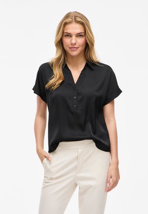 Blonde woman wearing a black short-sleeve buttoned blouse and light beige pants, standing with one hand in pocket against white background.