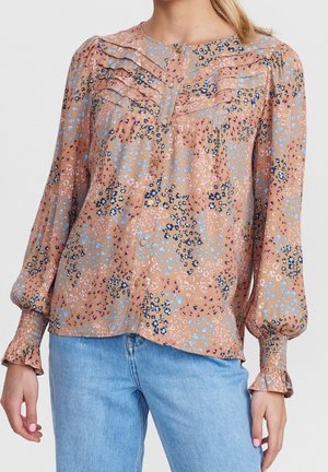 Ruusuinen pusero persikanvärisenä, jossa sinisiä, vaaleanpunaisia ja mustia kuvioita; siinä on napillinen edusta, laskostetut yksityiskohdat ja kuminauhalliset hihansuut.