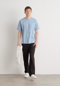 Lyseblå bomull T-skjorte, svarte bukser, hvite sneakers, holder en hvit genser, mot en enkel bakgrunn med jevn tekstur.