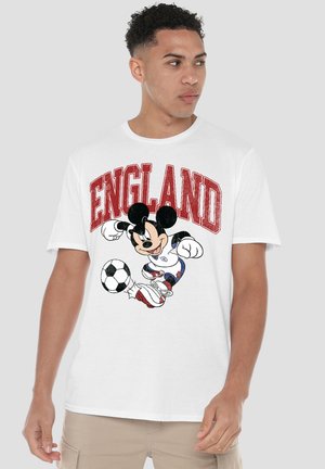Young man wearing white T-shirt with red "ENGLAND" text and Mickey Mouse kicking a soccer ball graphic, beige pants, looking right.