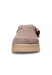 Scarpa in suede beige con punta arrotondata, accessori in metallo marrone e suola leggera a contrasto. Presenta una texture liscia e un design slip-on.