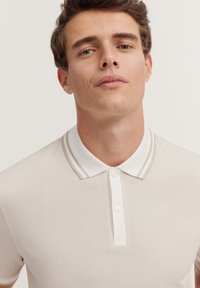 Polo-Shirt in hellem Beige mit strukturiertem Fischgrätmuster, weißem Kragen und doppelten Streifenakzenten. Mit Knopfleiste.