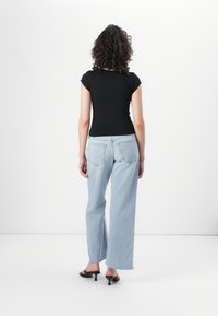 Juodas prigludęs marškinėliai, šviesiai mėlynos plačios kojų džinsai su suplėšytu kraštu ir juodos kulninės šlepetės, matoma iš nugaros ant neutralios fono.