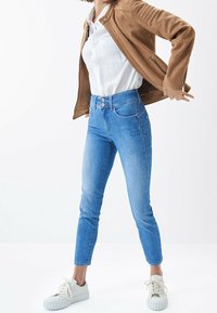 Světlé modré úzké džíny s vysokým pasem, dvěma předními kapsami a zapínáním na knoflíky, kombinované s hnědou semišovou bundou a bílými teniskami.