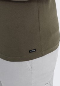 Olivgrünes Baumwoll-T-Shirt mit rundem Halsauschnitt und kurzen Ärmeln, mit einem kleinen schwarzen "Ombre"-Etikett am Saum. Weiße Hosen sind teilweise sichtbar.