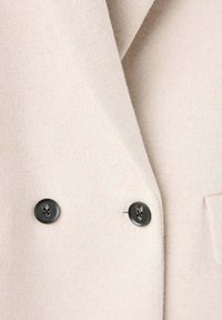 Gros plan d'un manteau en laine beige avec deux boutons noirs et une poche visible sur le côté droit.