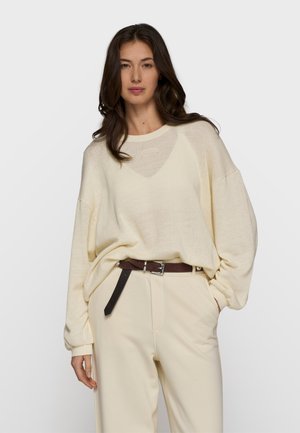 Kobieta ubrana w luźny kremowy sweter z dzianiny, kremowe spodnie z wysokim stanem i ciemnobrązowy pasek, stojąca z jedną ręką w kieszeni.