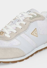 Baskets blanches et beiges avec lacets blancs, détails en daim et un triangle logo Guess doré sur le côté au-dessus d’une semelle en caoutchouc couleur gomme.