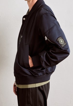 Homme portant une veste bomber noire avec des poignets côtelés, des poches latérales et une poche zippée sur la manche avec un emblème circulaire.