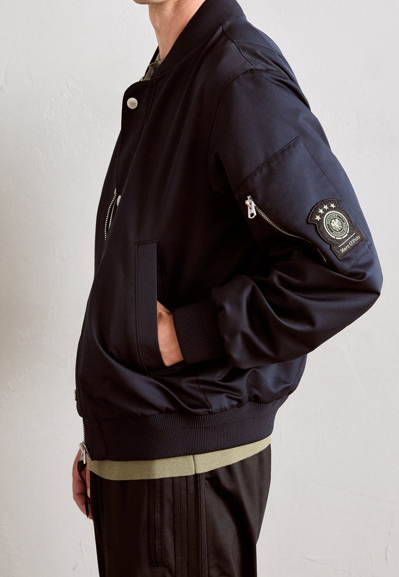 Homme portant une veste bomber noire avec des poignets côtelés, des poches latérales et une poche zippée sur la manche avec un emblème circulaire.
