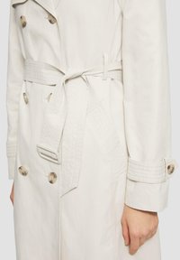 Jasnobeżowy płaszcz trencz z dwurzędowymi guzikami, paskiem wiązanym w talii oraz teksturowanymi detalami na rękawach i kieszeniach.