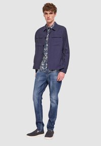 Giacca blu navy con chiusura a bottoni e due tasche sul petto sopra una camicia fantasia, abbinata a jeans blu e scarpe scure slip-on.
