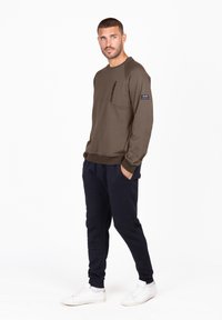 Sudadera verde oliva con cuello redondo, bolsillo en el pecho con cremallera y puños acanalados. Combinada con pantalones de chándal azul marino y zapatillas blancas.