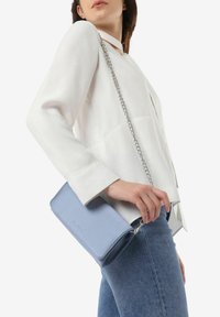 Borsa a tracolla in pelle azzurro chiaro con tracolla a catena, caratterizzata da una forma rettangolare elegante e logo impresso, tenuta da una donna che indossa una maglietta bianca.