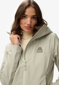 Lichtbeige waterdichte jas met capuchon, voorzien van een ritssluitingen en een logopatch op de borst. Glad textuur en minimalistisch ontwerp.