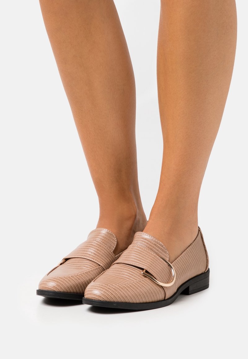 Jambes portant des mocassins beige texturés à enfiler avec un détail de boucle dorée et des talons bas noirs sur un fond clair uni.