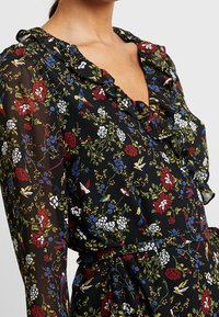 Blusa negra con flores que presenta un escote con volantes, patrones intrincados de flores y pájaros en rojo, blanco, azul y verde sobre una tela transparente.