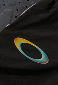Un tejido negro con una textura suave presenta un logotipo circular multicolor en tonos de azul, amarillo y rosa. El fondo muestra un material gris texturizado.