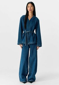 Tenue en denim composée d'un haut à manches longues, ceinturé, avec un décolleté en V profond et un pantalon à jambes larges. Couleur bleu foncé avec une texture et des coutures denim classiques.