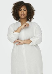 Camicia bianca in lino con colletto alla mandarino, maniche lunghe arrotolate ai polsini, caratterizzata da una vestibilità rilassata e superficie texturizzata.