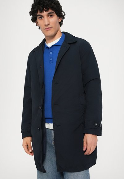 Mørkeblå vandafvisende frakke med knapper, en spids krave og sidelommer, parret med en blå poloshirt og denimjeans.