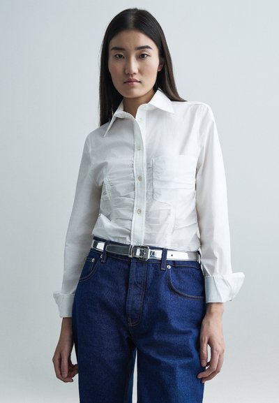 Jauna moteris su ilgais tamsiais plaukais, dėvinti baltą raukinį mygtukų marškinių, tamsiai mėlynus džinsus ir blizgantį sidabrinį diržą, stovinti prieš neutralų fono.