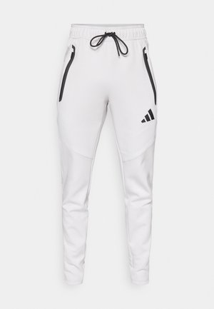 White athletic pants with a slim fit. Features black accents, zippered pockets, and an elastic drawstring waistband. Adidas logo displayed.