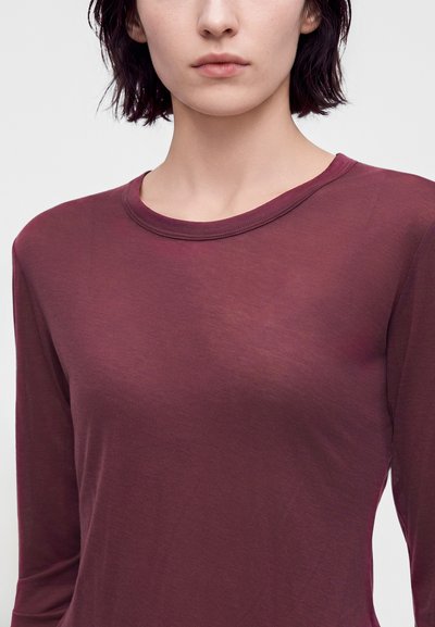Femme aux cheveux courts et foncés portant une chemise ajustée à manches longues bordeaux sur un fond clair uni.