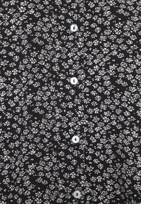 Chemise en tissu noir avec un motif floral blanc, petites boutons sur le devant et une surface texturée.
