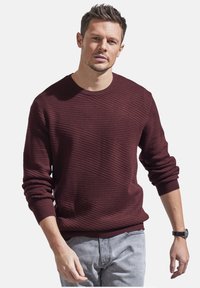 Burgunderfarbener Rundhals-Pullover mit diagonaler Rippstruktur, langen Ärmeln und gerippten Bündchen. Getragen über hellgrauen Hosen.